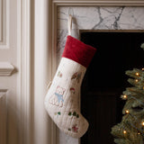 A cream-colored Christmas stocking with embroidered teddy bear motifs and a red velvet cuff, hanging in front of a marble fireplace next to a lit Christmas tree.
