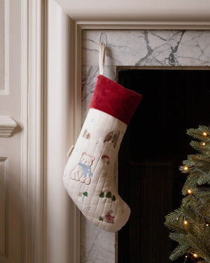 A cream-colored Christmas stocking with embroidered teddy bear motifs and a red velvet cuff, hanging in front of a marble fireplace next to a lit Christmas tree.