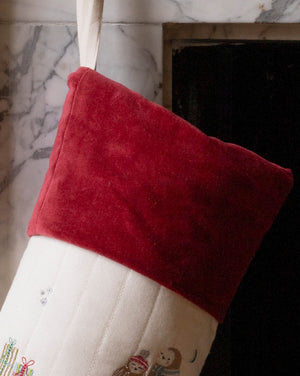 A Christmas stocking with a red velvet cuff and a white body decorated with small holiday illustrations, hanging in front of a fireplace.