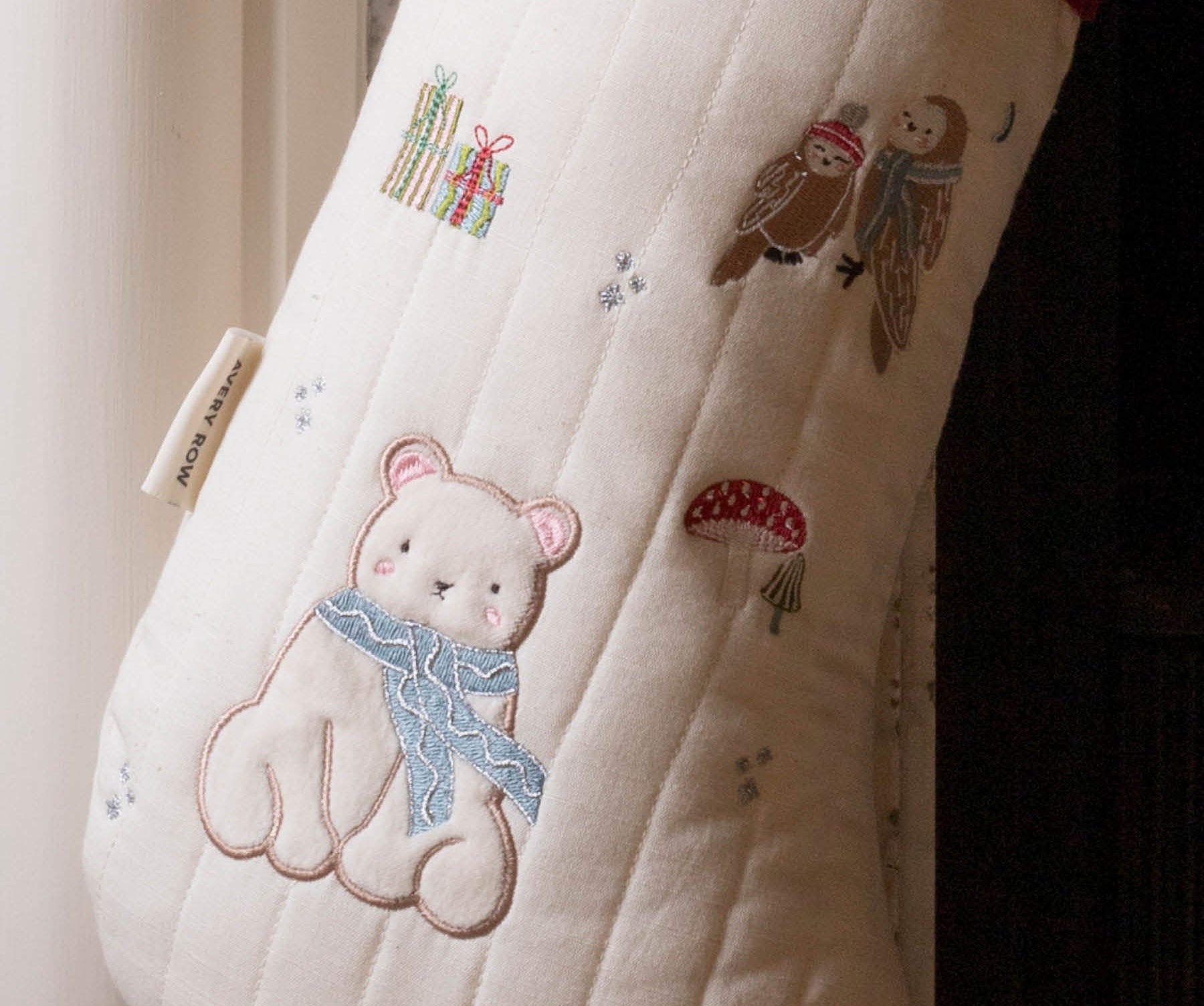 A close-up of a quilted Christmas stocking in cream with embroidered woodland motifs, including a teddy bear in a blue scarf, an owl, gifts, mushrooms, and trees. A red cuff is visible at the top, and a small fabric tag is attached near the left side.