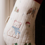 A close-up of a quilted Christmas stocking in cream with embroidered woodland motifs, including a teddy bear in a blue scarf, an owl, gifts, mushrooms, and trees. A red cuff is visible at the top, and a small fabric tag is attached near the left side.