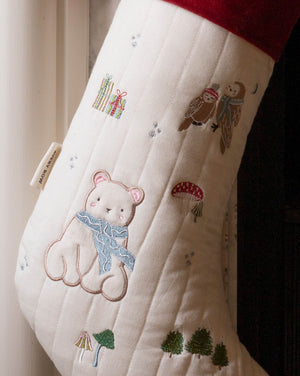 A close-up of a quilted Christmas stocking in cream with embroidered woodland motifs, including a teddy bear in a blue scarf, an owl, gifts, mushrooms, and trees. A red cuff is visible at the top, and a small fabric tag is attached near the left side.