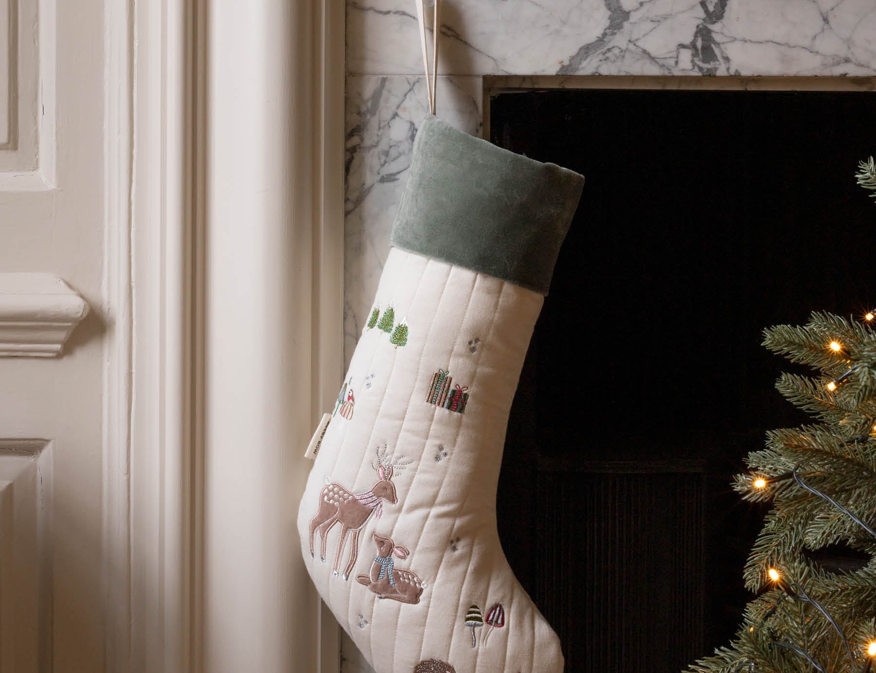 A quilted Christmas stocking with a cream body, green velvet cuff, and embroidered woodland motifs including a deer, hanging by a hook in a fireplace next to a lit Christmas tree.