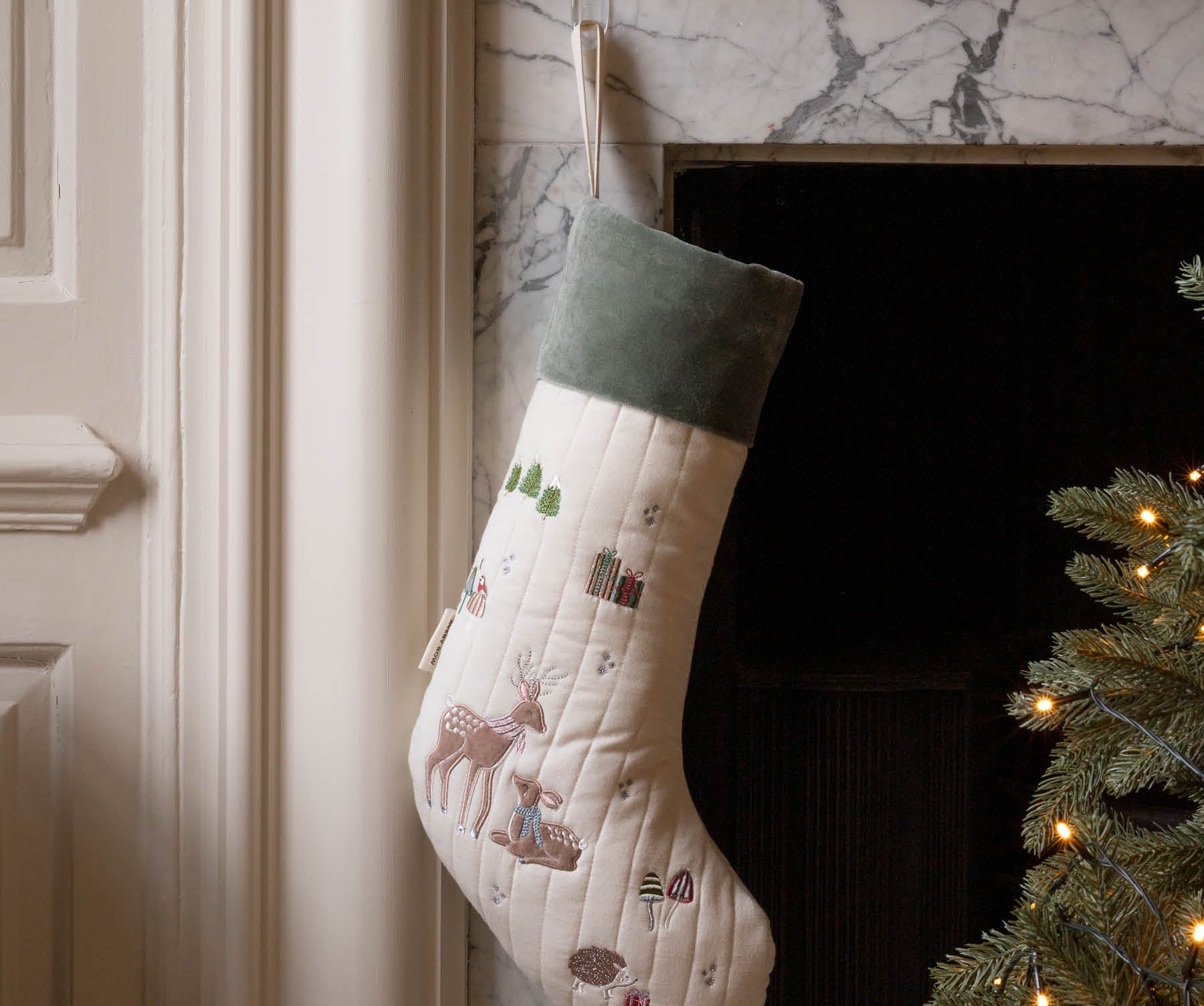 A quilted Christmas stocking with a cream body, green velvet cuff, and embroidered woodland motifs including a deer, hanging by a hook in a fireplace next to a lit Christmas tree.