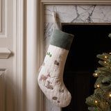 A quilted Christmas stocking with a cream body, green velvet cuff, and embroidered woodland motifs including a deer, hanging by a hook in a fireplace next to a lit Christmas tree.