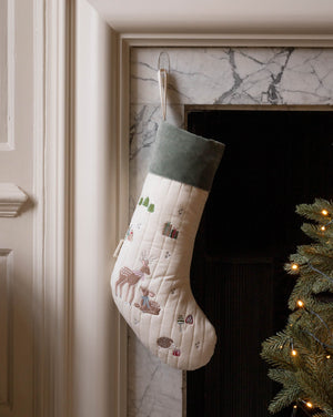 A quilted Christmas stocking with a cream body, green velvet cuff, and embroidered woodland motifs including a deer, hanging by a hook in a fireplace next to a lit Christmas tree.