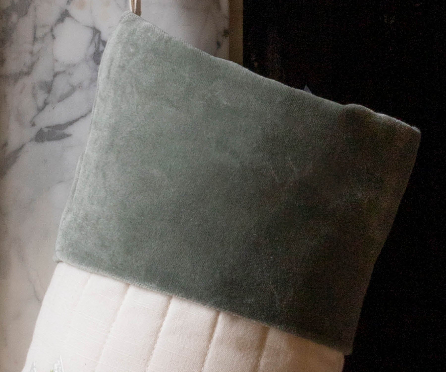 A Christmas stocking made of sage green velvet and cream quilted fabric, hanging in front of a marble fireplace.