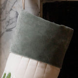 A Christmas stocking made of sage green velvet and cream quilted fabric, hanging in front of a marble fireplace.