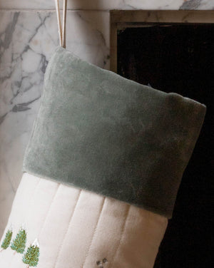 A Christmas stocking made of sage green velvet and cream quilted fabric, hanging in front of a marble fireplace.