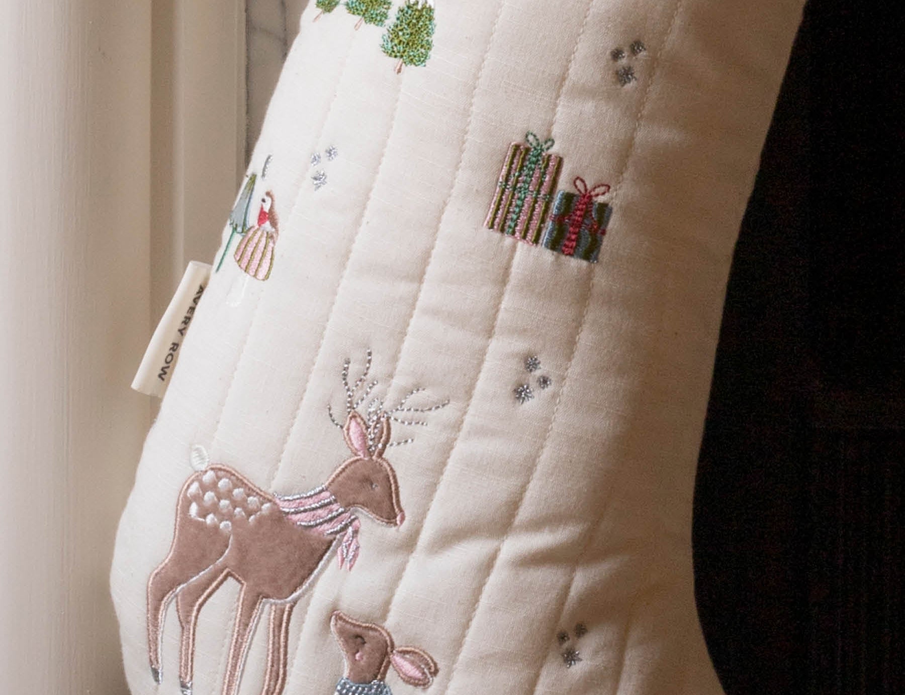 An off-white quilted Christmas stocking with embroidered deer, gifts, trees, and small snowflake details, shown hanging near a window.
