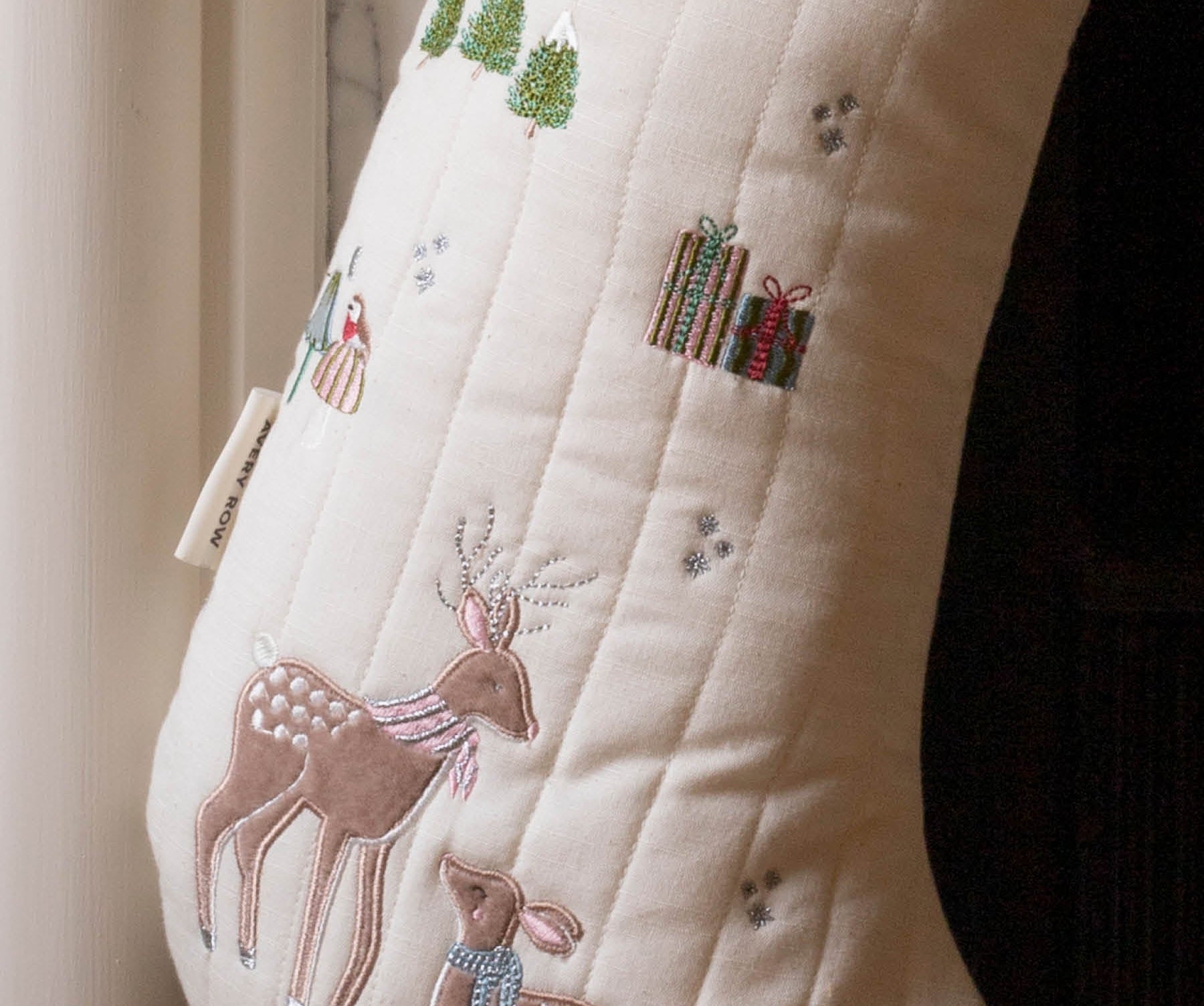 An off-white quilted Christmas stocking with embroidered deer, gifts, trees, and small snowflake details, shown hanging near a window.