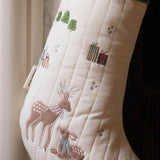 An off-white quilted Christmas stocking with embroidered deer, gifts, trees, and small snowflake details, shown hanging near a window.