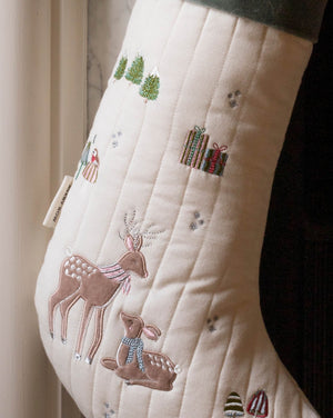 An off-white quilted Christmas stocking with embroidered deer, gifts, trees, and small snowflake details, shown hanging near a window.