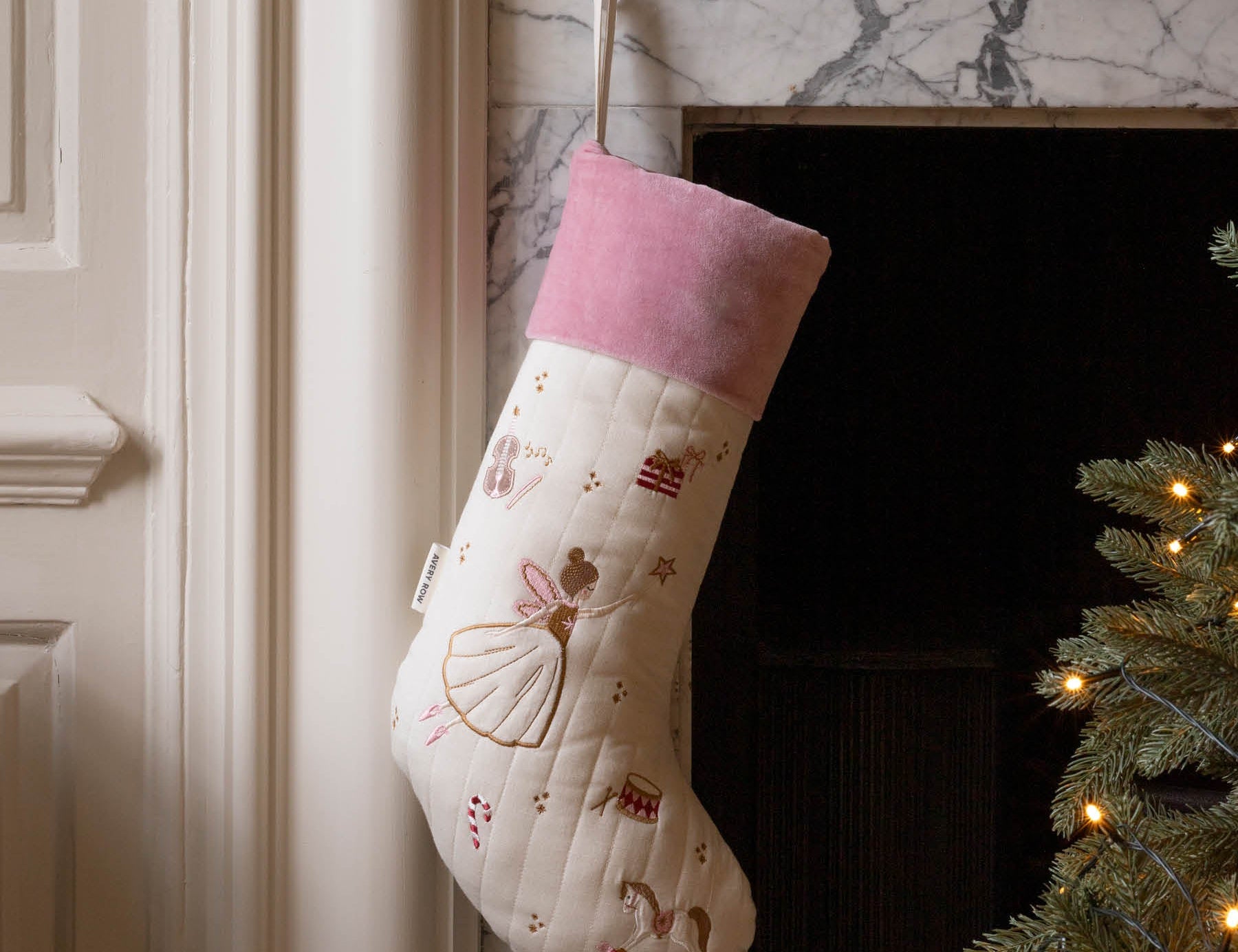 A cream quilted Christmas stocking with a pink velvet cuff, decorated with embroidered ballerina and candy cane motifs, hanging on a marble fireplace hearth next to a lit Christmas tree.