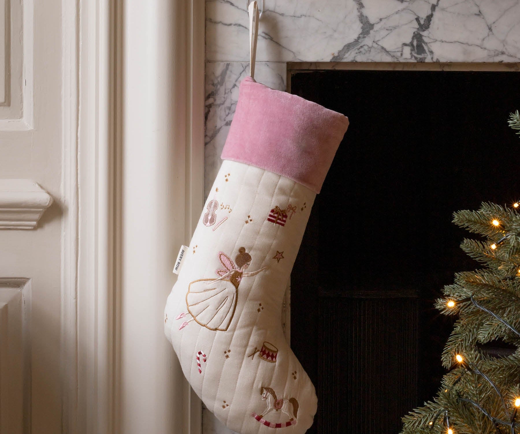A cream quilted Christmas stocking with a pink velvet cuff, decorated with embroidered ballerina and candy cane motifs, hanging on a marble fireplace hearth next to a lit Christmas tree.