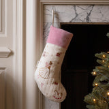 A cream quilted Christmas stocking with a pink velvet cuff, decorated with embroidered ballerina and candy cane motifs, hanging on a marble fireplace hearth next to a lit Christmas tree.