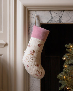 A cream quilted Christmas stocking with a pink velvet cuff, decorated with embroidered ballerina and candy cane motifs, hanging on a marble fireplace hearth next to a lit Christmas tree.