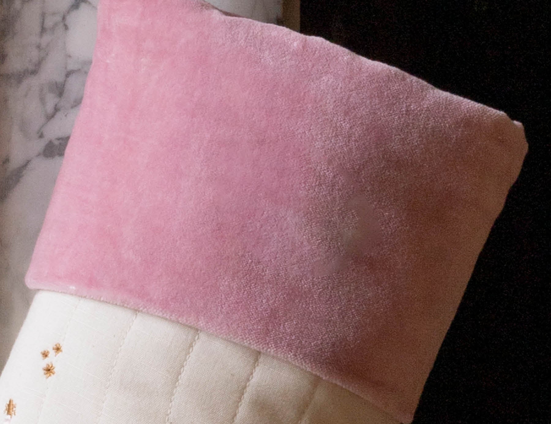 A close-up of a Christmas stocking with a soft pink velvet cuff and a cream quilted body decorated with embroidered gold stars and a small red gift motif, hanging in front of a fireplace.