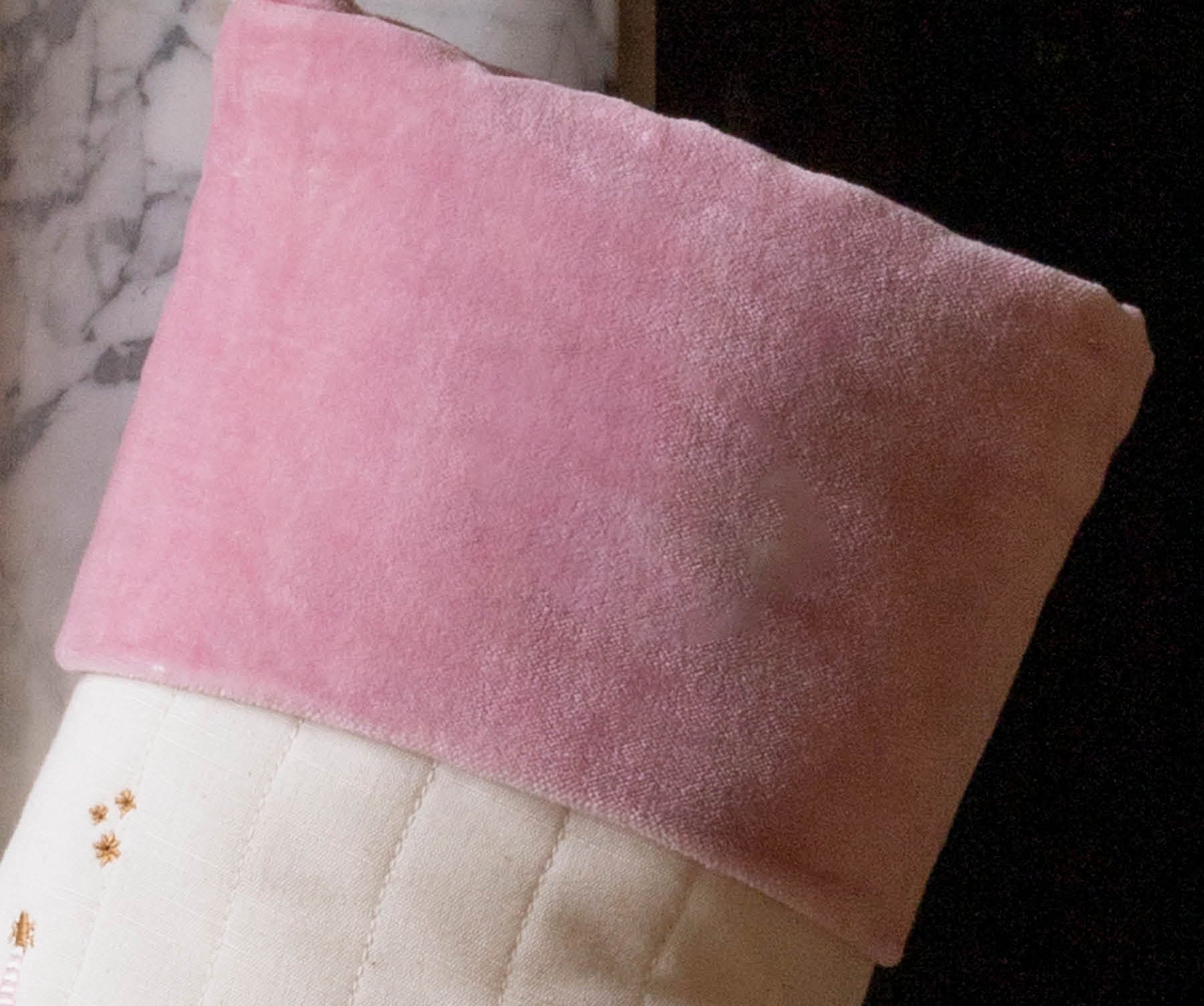 A close-up of a Christmas stocking with a soft pink velvet cuff and a cream quilted body decorated with embroidered gold stars and a small red gift motif, hanging in front of a fireplace.