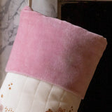 A close-up of a Christmas stocking with a soft pink velvet cuff and a cream quilted body decorated with embroidered gold stars and a small red gift motif, hanging in front of a fireplace.