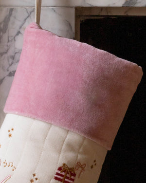 A close-up of a Christmas stocking with a soft pink velvet cuff and a cream quilted body decorated with embroidered gold stars and a small red gift motif, hanging in front of a fireplace.
