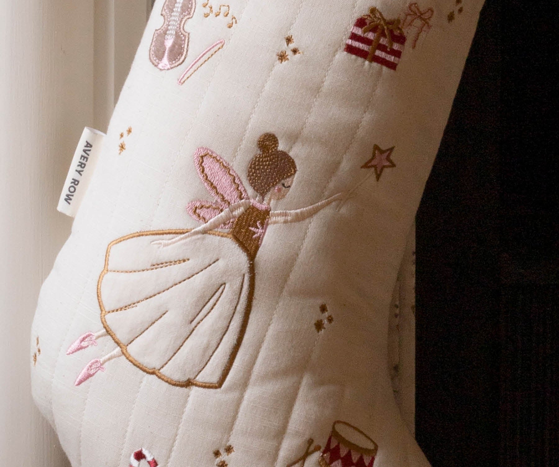 An off-white quilted Christmas stocking with embroidered festive motifs including a ballerina, stars, a violin, a gift box, and candy canes. The stocking features a pink ribbed cuff at the top and a white tag on the side that reads 'A NEW WAY'.