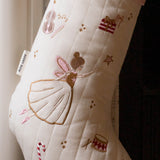 An off-white quilted Christmas stocking with embroidered festive motifs including a ballerina, stars, a violin, a gift box, and candy canes. The stocking features a pink ribbed cuff at the top and a white tag on the side that reads 'A NEW WAY'.
