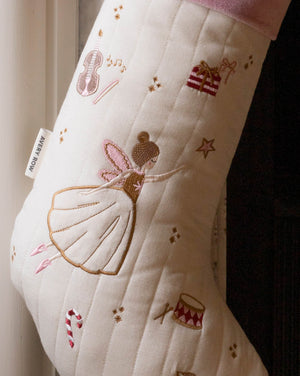 An off-white quilted Christmas stocking with embroidered festive motifs including a ballerina, stars, a violin, a gift box, and candy canes. The stocking features a pink ribbed cuff at the top and a white tag on the side that reads 'A NEW WAY'.