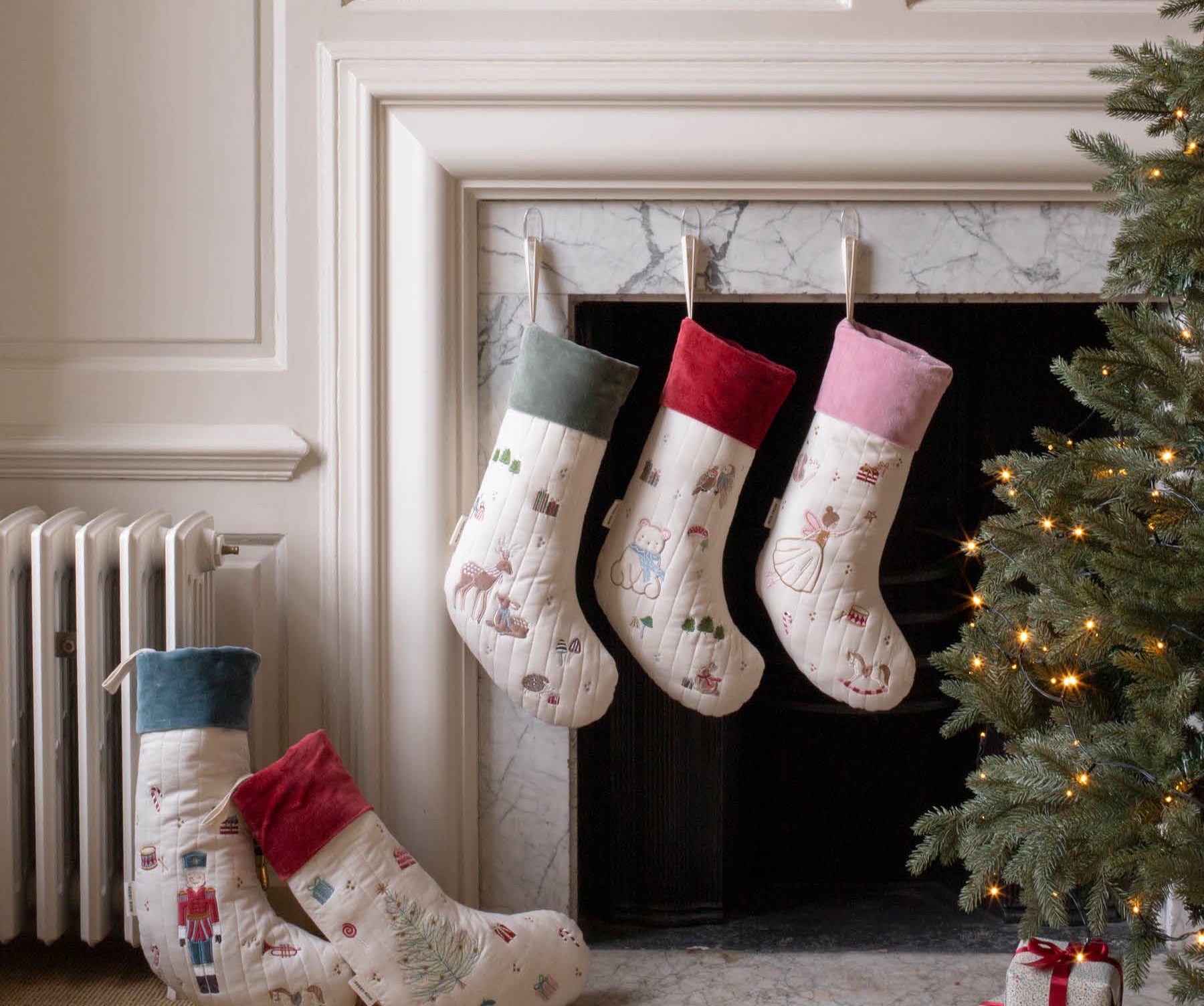 Four Christmas stockings with festive prints are displayed around a fireplace. Three stockings hang from the mantel, while two more are placed on the floor beside a lit Christmas tree and wrapped gift boxes.