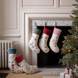 Four Christmas stockings with festive prints are displayed around a fireplace. Three stockings hang from the mantel, while two more are placed on the floor beside a lit Christmas tree and wrapped gift boxes.
