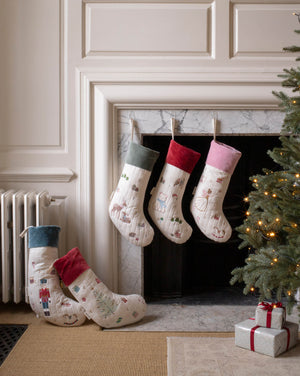 Four Christmas stockings with festive prints are displayed around a fireplace. Three stockings hang from the mantel, while two more are placed on the floor beside a lit Christmas tree and wrapped gift boxes.