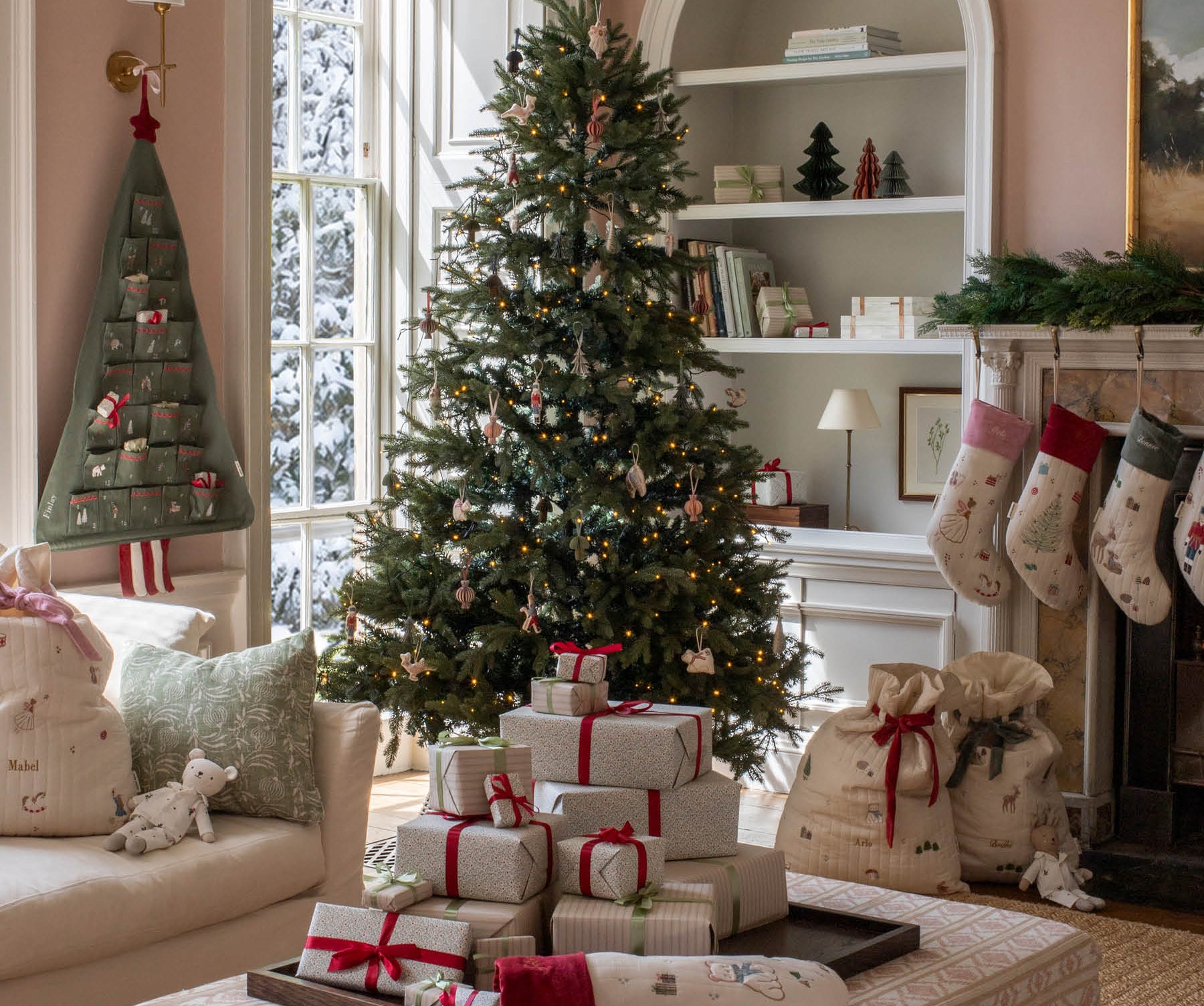 A beautifully decorated Christmas living room featuring a tall evergreen tree with lights and ornaments, wrapped gift boxes on a patterned ottoman, stockings hanging from a fireplace, and burlap Santa bags beside it.