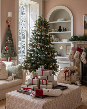 A beautifully decorated Christmas living room featuring a tall evergreen tree with lights and ornaments, wrapped gift boxes on a patterned ottoman, stockings hanging from a fireplace, and burlap Santa bags beside it.