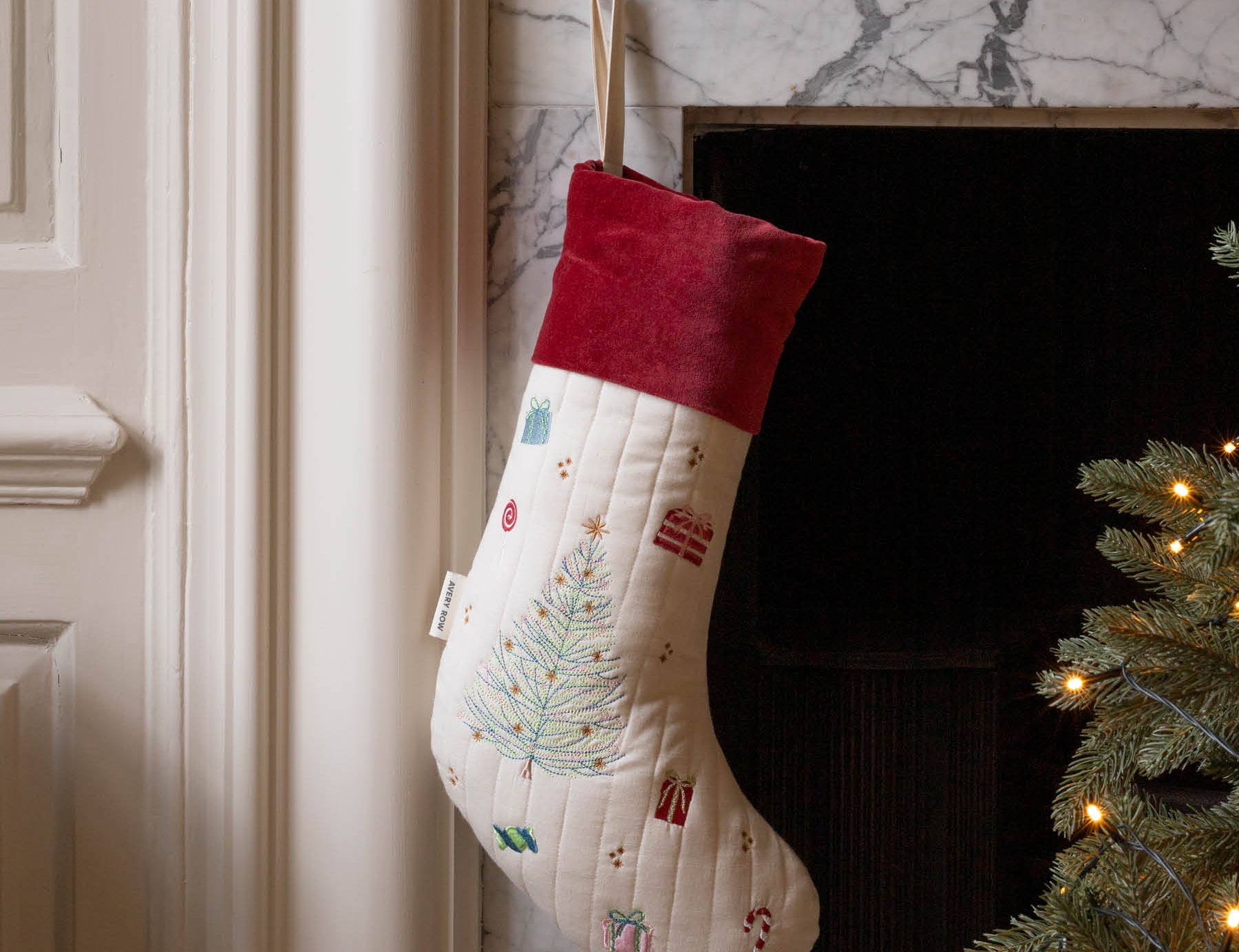 A cream quilted Christmas stocking with a red velvet cuff, decorated with a light blue Christmas tree and small holiday motifs, hanging in front of a fireplace next to a lit Christmas tree.