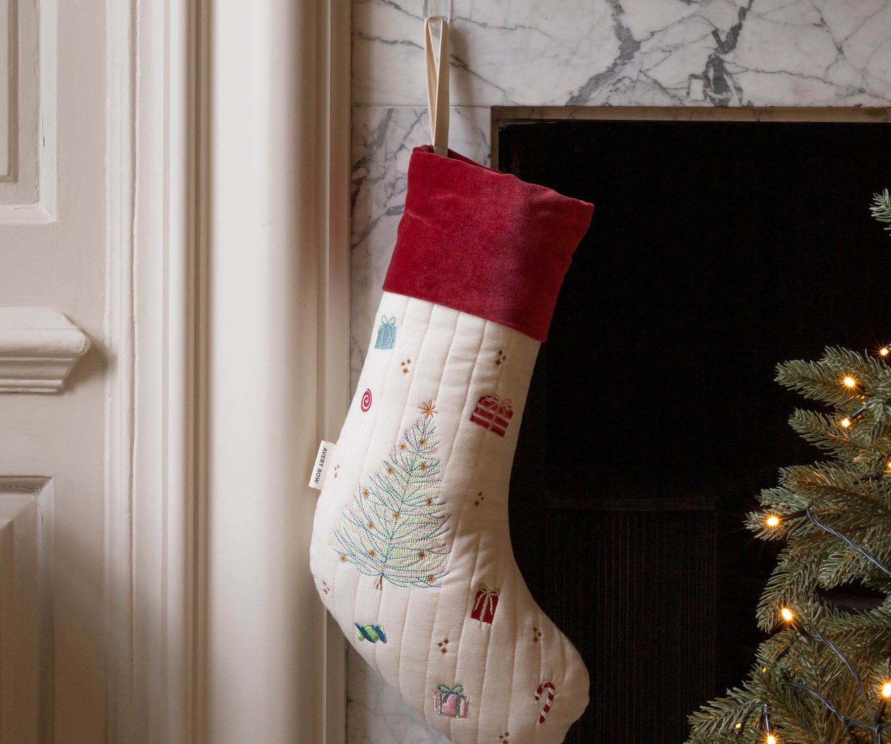 A cream quilted Christmas stocking with a red velvet cuff, decorated with a light blue Christmas tree and small holiday motifs, hanging in front of a fireplace next to a lit Christmas tree.