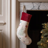 A cream quilted Christmas stocking with a red velvet cuff, decorated with a light blue Christmas tree and small holiday motifs, hanging in front of a fireplace next to a lit Christmas tree.