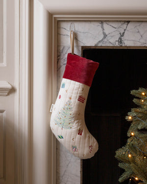 A cream quilted Christmas stocking with a red velvet cuff, decorated with a light blue Christmas tree and small holiday motifs, hanging in front of a fireplace next to a lit Christmas tree.