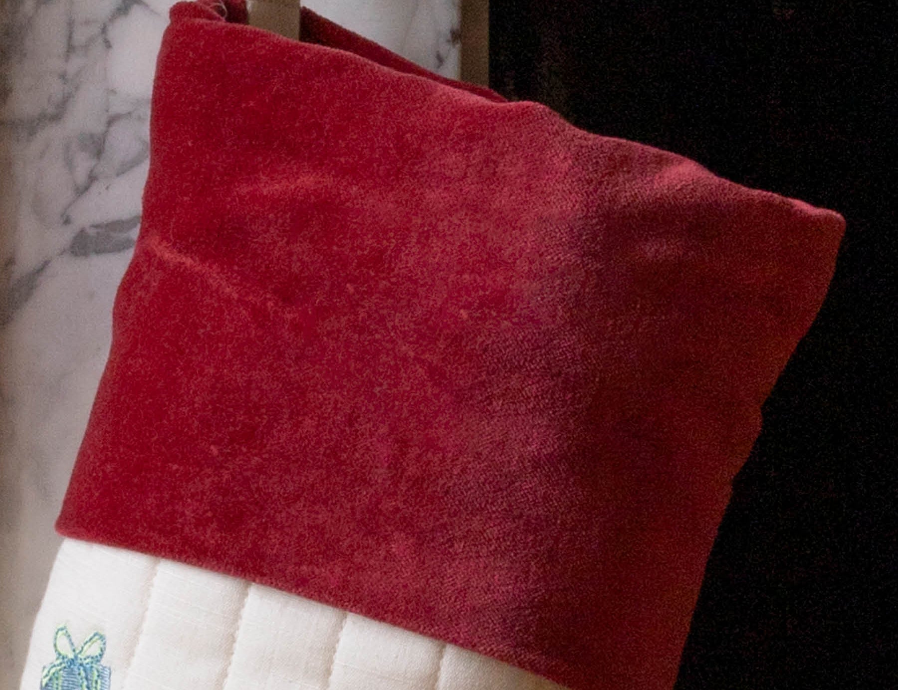 A close-up of a Christmas stocking with a deep red velvet cuff and a white quilted body, hanging in front of a fireplace.