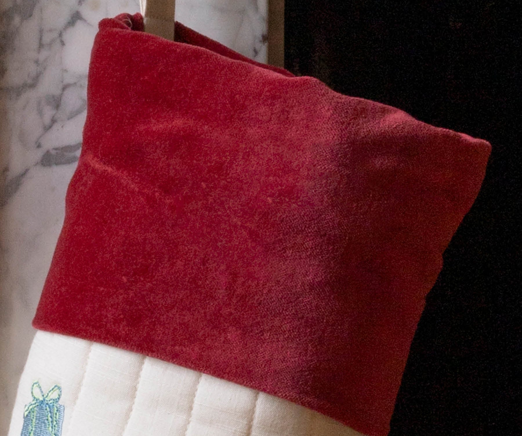 A close-up of a Christmas stocking with a deep red velvet cuff and a white quilted body, hanging in front of a fireplace.