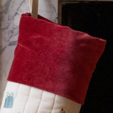 A close-up of a Christmas stocking with a deep red velvet cuff and a white quilted body, hanging in front of a fireplace.