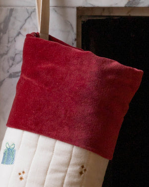 A close-up of a Christmas stocking with a deep red velvet cuff and a white quilted body, hanging in front of a fireplace.