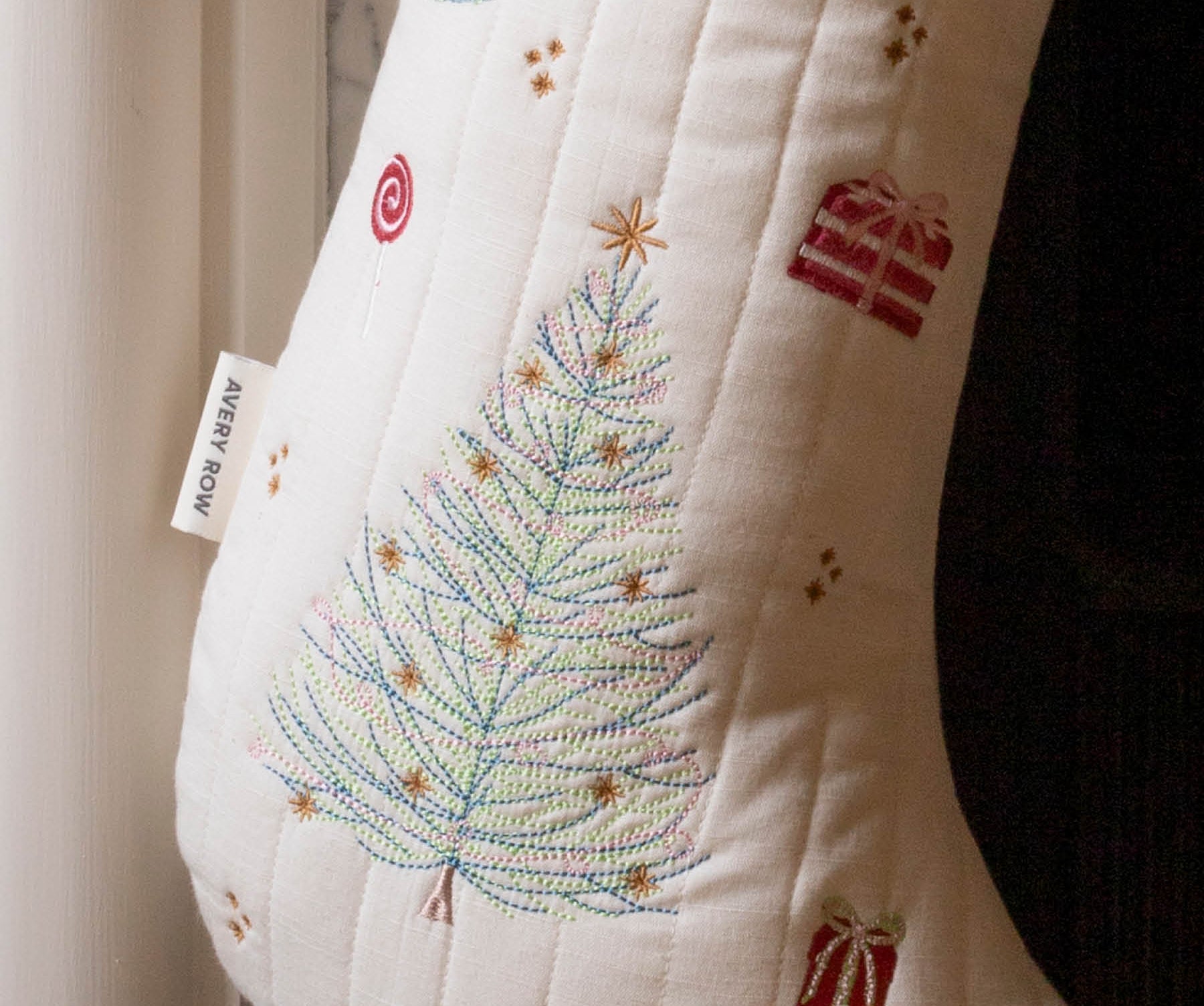 An off-white quilted Christmas stocking with a colorful embroidered tree, gift boxes, and a red cuff, hanging next to a white curtain.