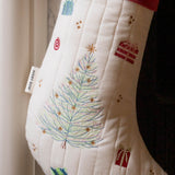 An off-white quilted Christmas stocking with a colorful embroidered tree, gift boxes, and a red cuff, hanging next to a white curtain.