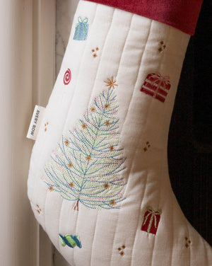An off-white quilted Christmas stocking with a colorful embroidered tree, gift boxes, and a red cuff, hanging next to a white curtain.