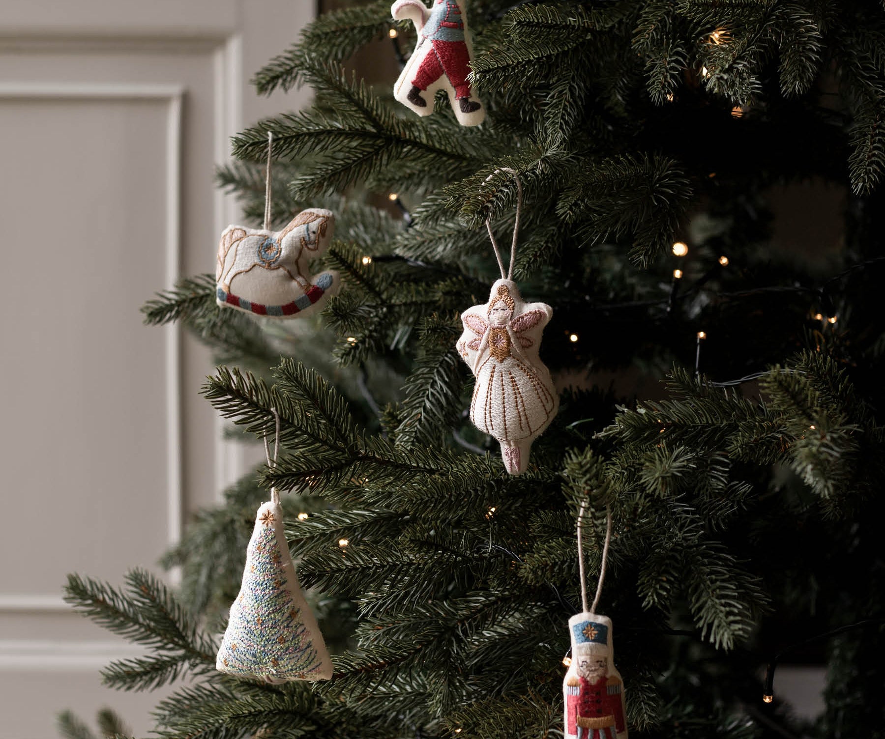 Handmade knitted Christmas ornaments hanging on a decorated evergreen tree with warm string lights. The ornaments include a rocking horse, a angel, a nutcracker, a Christmas tree, and a small figure in colorful yarn.