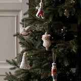 Handmade knitted Christmas ornaments hanging on a decorated evergreen tree with warm string lights. The ornaments include a rocking horse, a angel, a nutcracker, a Christmas tree, and a small figure in colorful yarn.