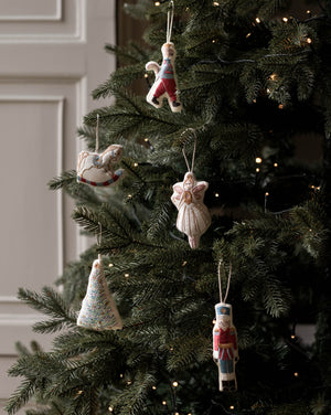 Handmade knitted Christmas ornaments hanging on a decorated evergreen tree with warm string lights. The ornaments include a rocking horse, a angel, a nutcracker, a Christmas tree, and a small figure in colorful yarn.