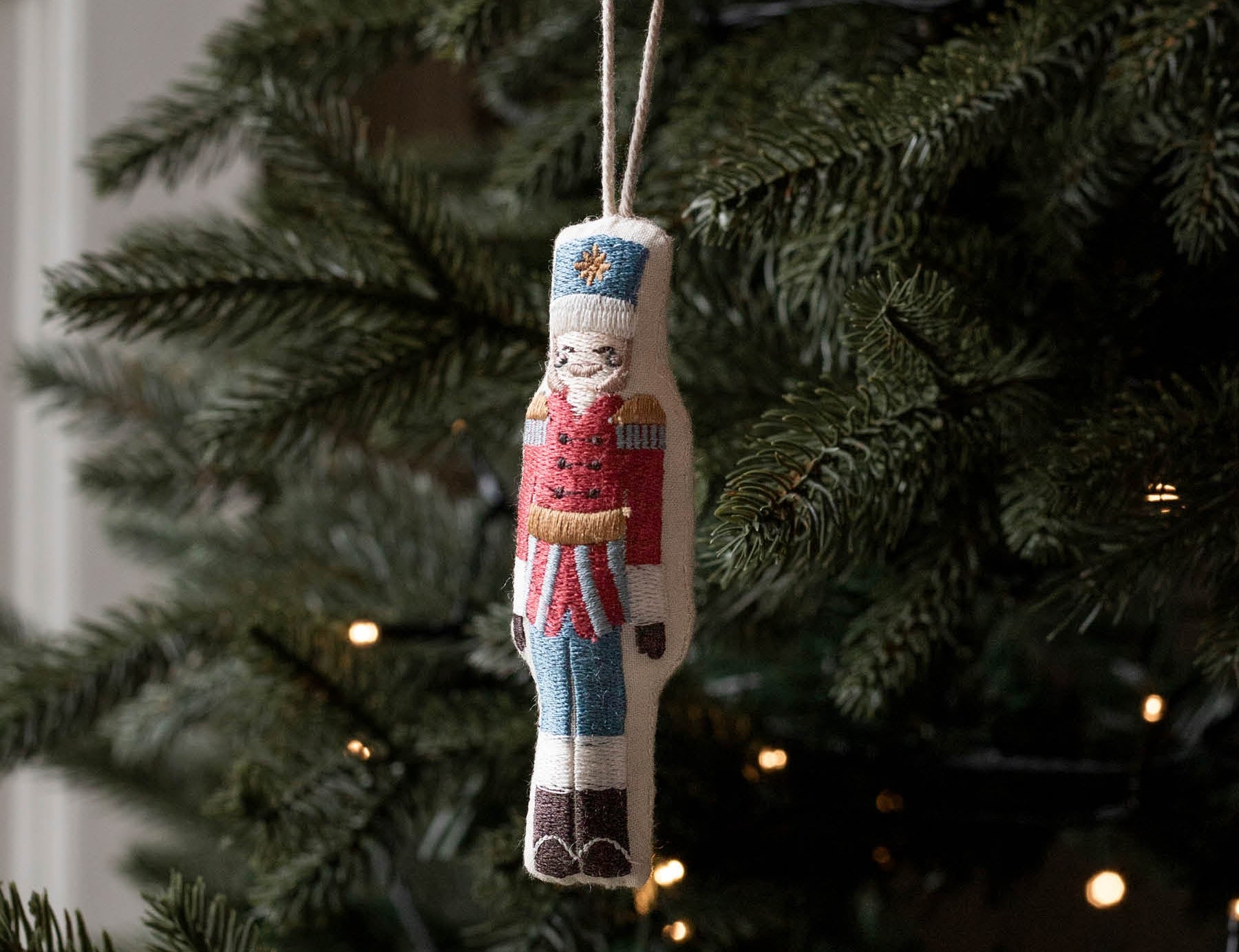 A hand holds a small embroidered nutcracker Christmas ornament against the green branches of a lit Christmas tree.