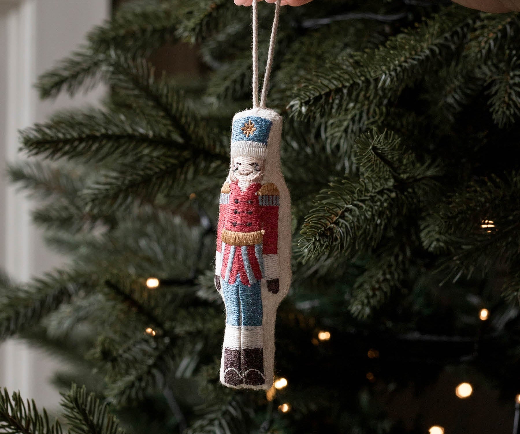 A hand holds a small embroidered nutcracker Christmas ornament against the green branches of a lit Christmas tree.