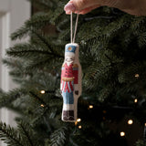 A hand holds a small embroidered nutcracker Christmas ornament against the green branches of a lit Christmas tree.